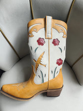 Pair of white cowboy boots with yellow accents and floral designs on a light gray surface.