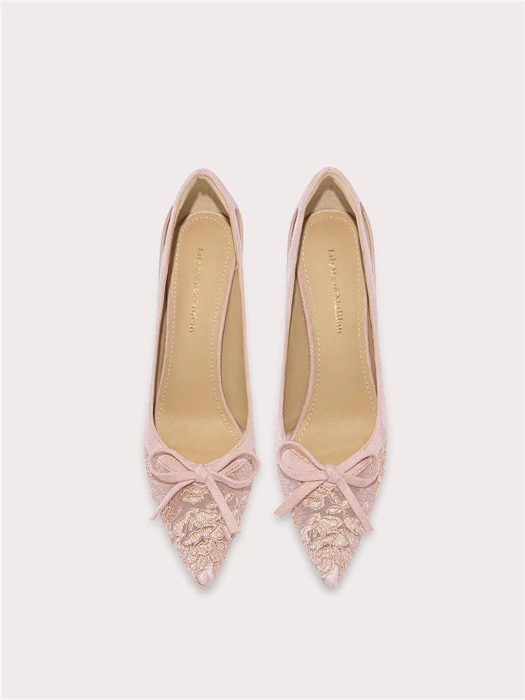 Pair of pink pointed-toe shoes with lace details and bow accents on a light gray background.
