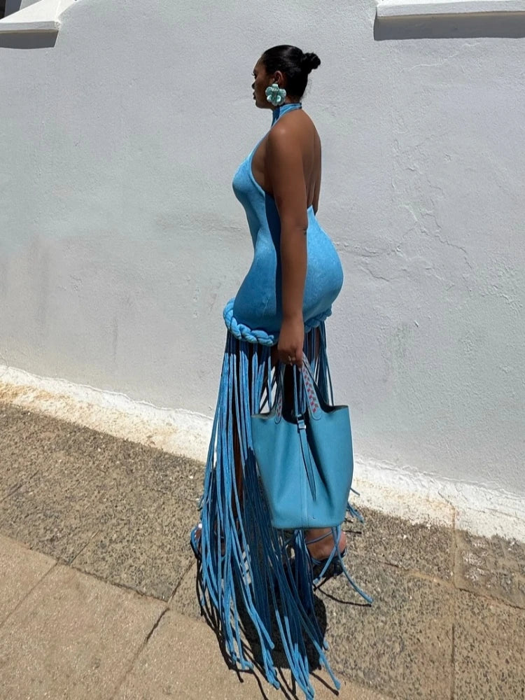 Woman in a blue dress with fringe holding a matching blue handbag against a gray wall.