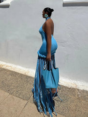 Woman in a blue dress with fringe holding a matching blue handbag against a gray wall.