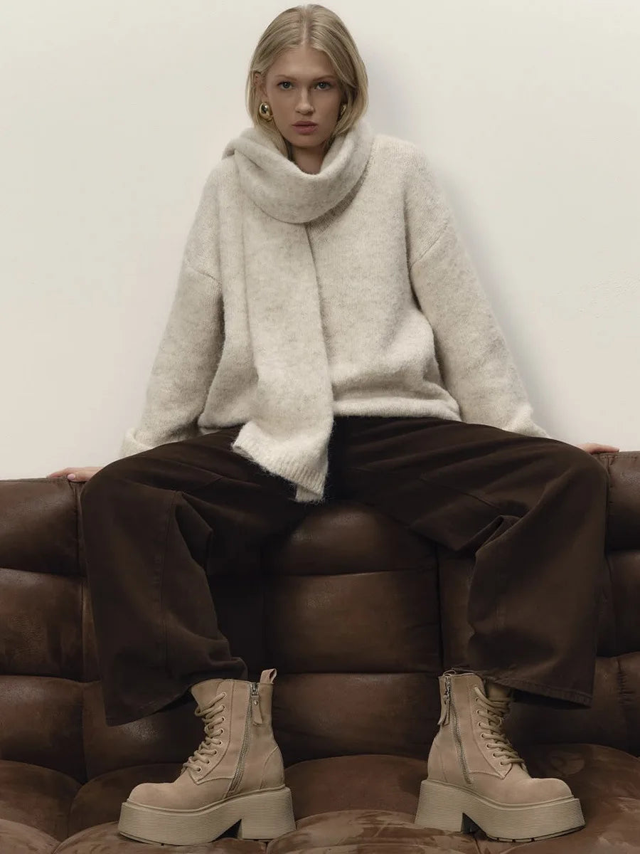 Person wearing a white sweater, scarf, and brown pants sitting on a brown couch.