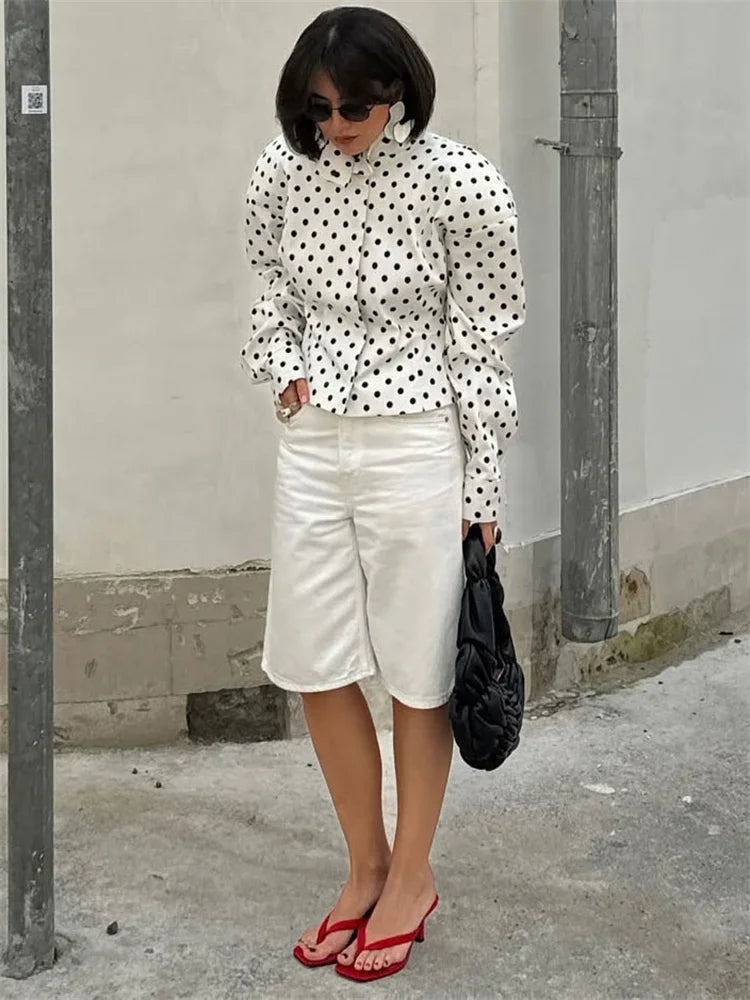 Woman wearing a polka dot blouse and white shorts standing against a plain wall.