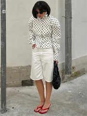 Woman wearing a polka dot blouse and white shorts standing against a plain wall.