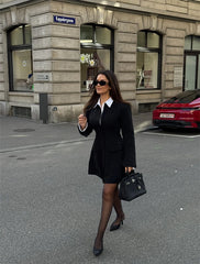 Woman in a black suit walking on a city street with a building and car in the background.