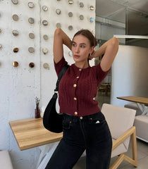 Woman posing in a modern indoor setting wearing a maroon knitted cardigan and black jeans.