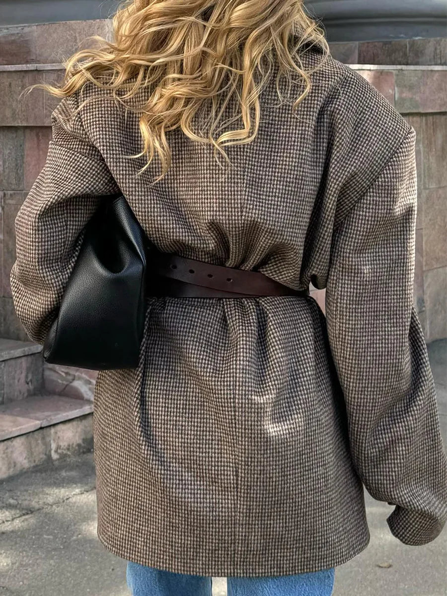 Person wearing a brown checkered coat with a black bag over their shoulder, standing against a stone wall.