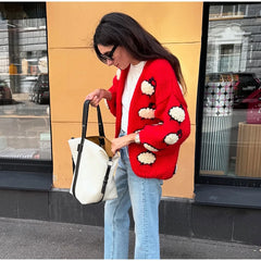 Woman in a red cardigan with white floral patterns holding a white bag against a yellow wall.