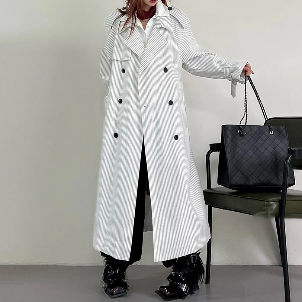 Person wearing a long white coat with black buttons, holding a black handbag against a neutral background.