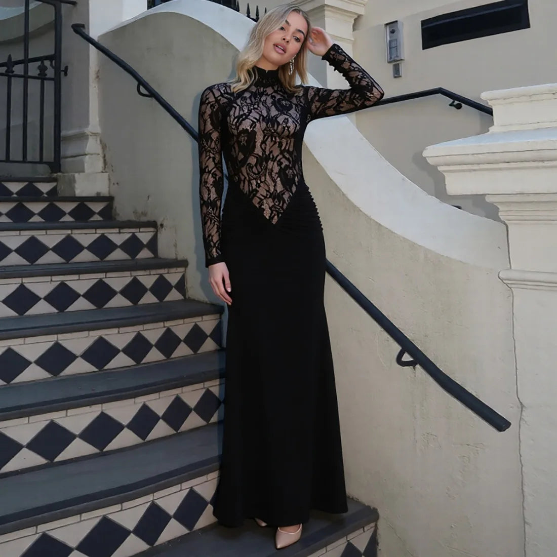 Woman in a black lace dress standing on a staircase with a white wall and black railing.