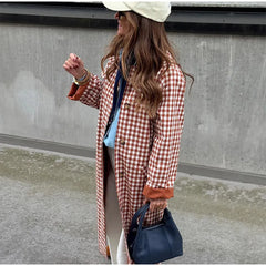 Woman wearing a checkered coat and holding a blue bag against a concrete wall.
