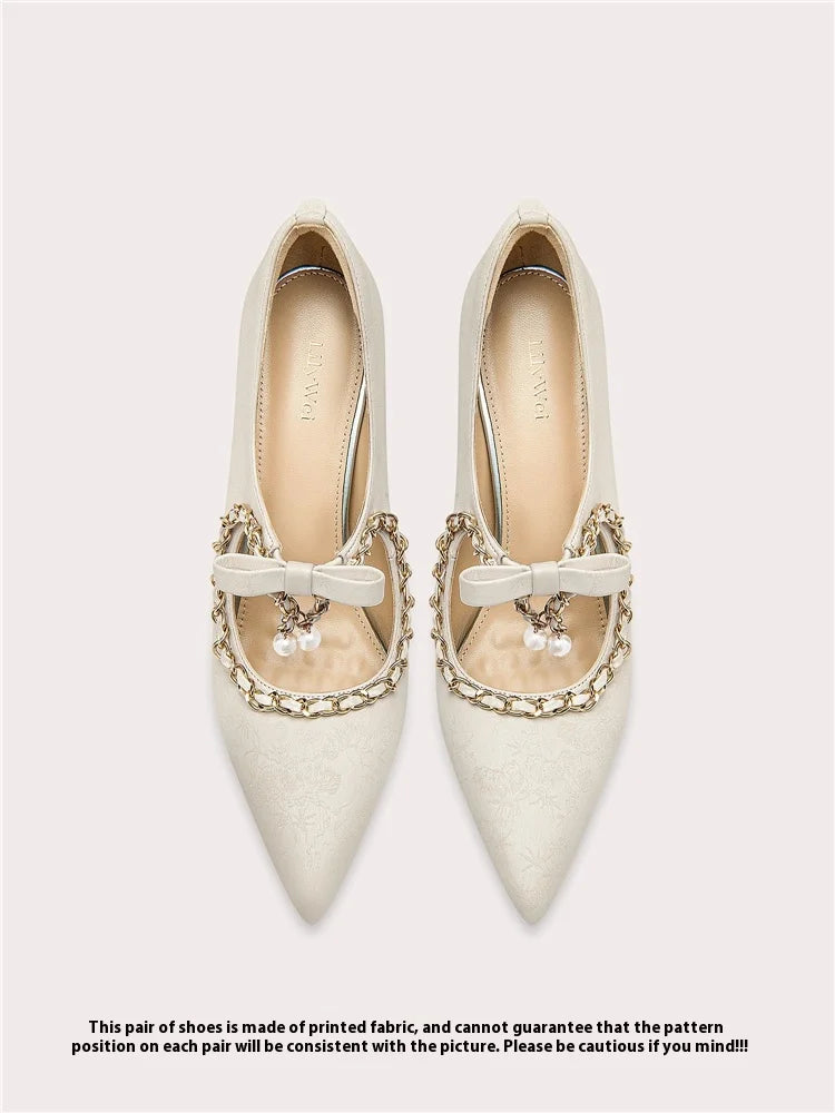 Pair of white pointed-toe shoes with decorative bows and chains on a light gray background.