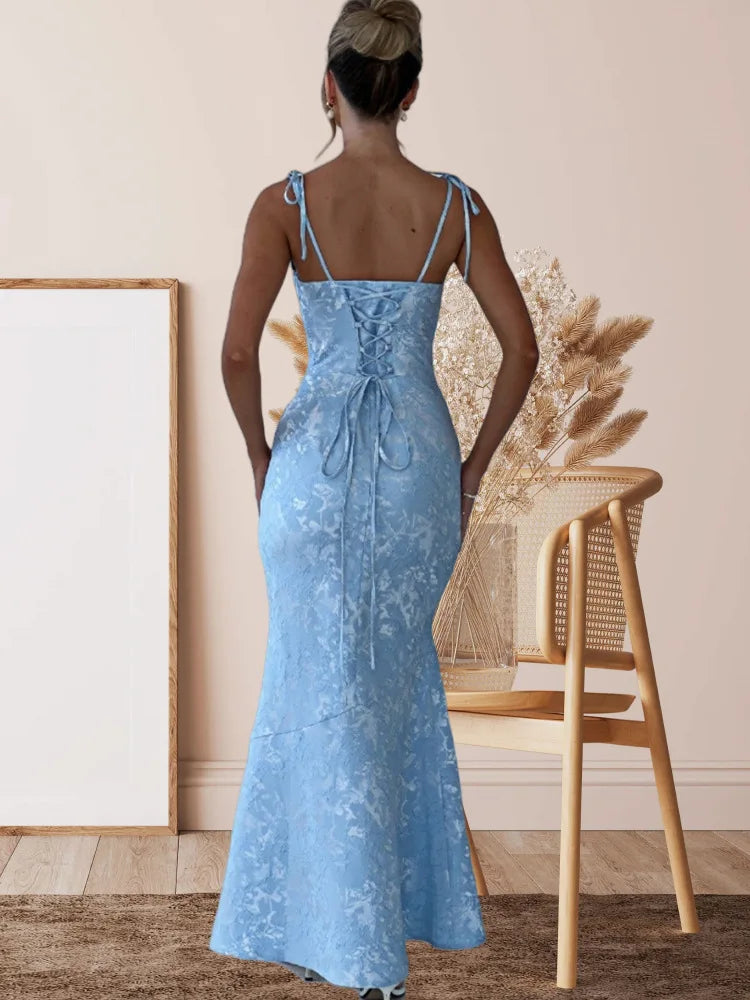 Woman wearing a blue lace dress standing in a room with a chair and decorative plants.