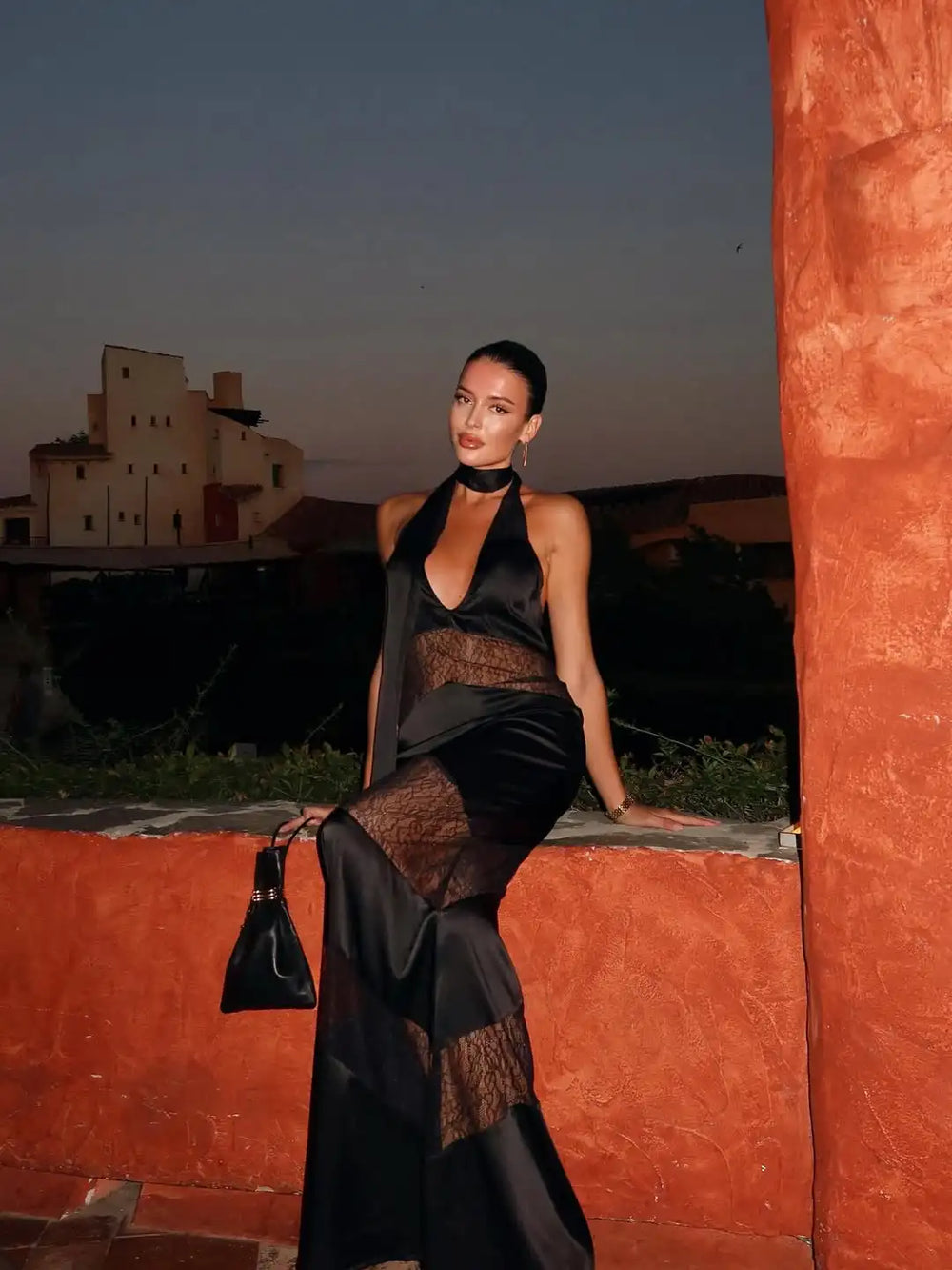 Woman in a black dress standing against an orange wall with a sunset or sunrise sky.