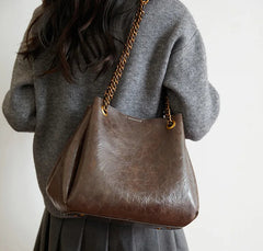 Brown leather handbag with gold chain strap worn by a person in a gray sweater.