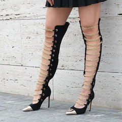 Black high-heeled boots with gold chain detailing worn by a person against a light gray wall.