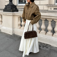 Person wearing a brown jacket and white skirt with a brown handbag, standing in front of a stone building.