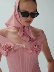 Woman wearing a pink dress with floral details and a matching headscarf against a plain background