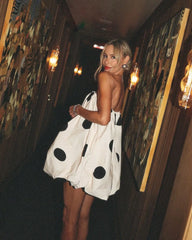 Woman in a polka dot dress standing in a dimly lit room with artistic wall decorations.