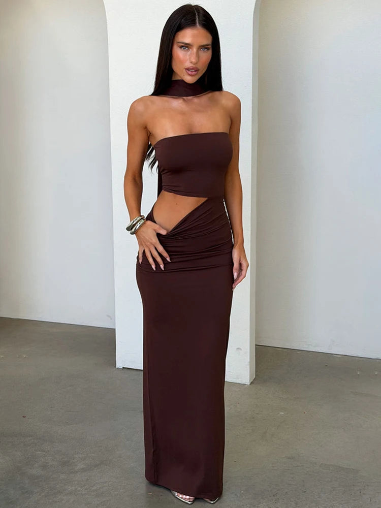 Woman wearing a brown strapless maxi dress with a side cutout and halter neck detail, posing against a white wall.