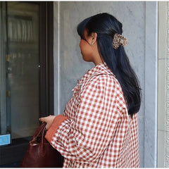 Woman in a red and white checkered shirt with a brown handbag, standing against a gray wall.