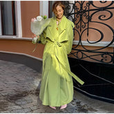 Woman in a bright green trench coat holding flowers, standing on a street with decorative iron railing.