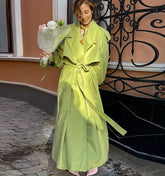 Woman in a bright green trench coat holding flowers, standing on a street with decorative iron railing.