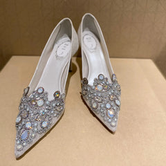Pair of white high-heeled shoes with silver embellishments on a beige surface.