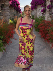 Woman in a floral dress standing in a garden with flowers and stone walls.