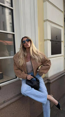 A woman in a khaki coat and blue jeans standing in front of a building entrance.