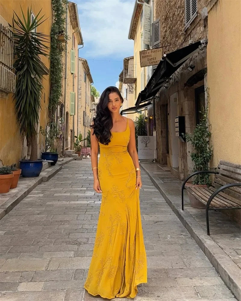 Woman in a yellow dress standing on a sunlit street with buildings and plants.