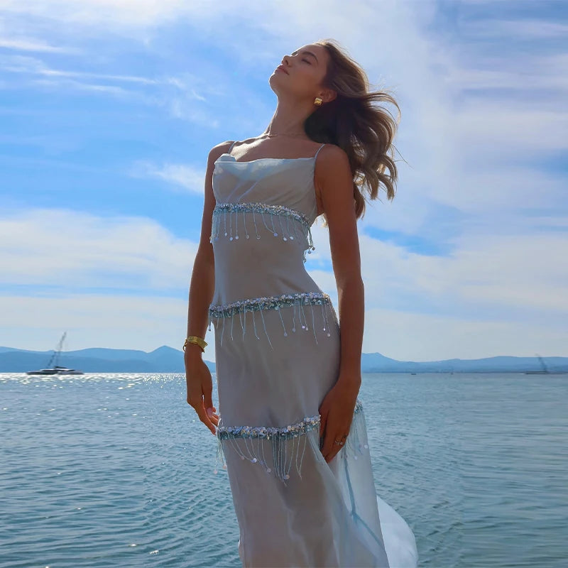 Woman in a white dress with beaded details standing by the water under a blue sky.