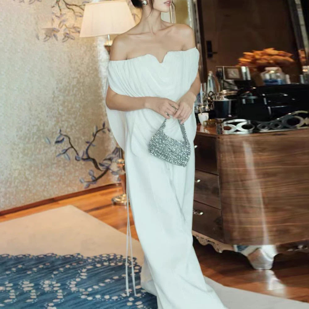 Woman in a white off-shoulder dress holding a silver clutch in a room with wooden furniture and decorative elements.