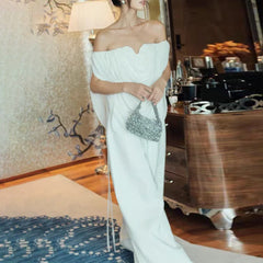 Woman in a white off-shoulder dress holding a silver clutch in a room with wooden furniture and decorative elements.