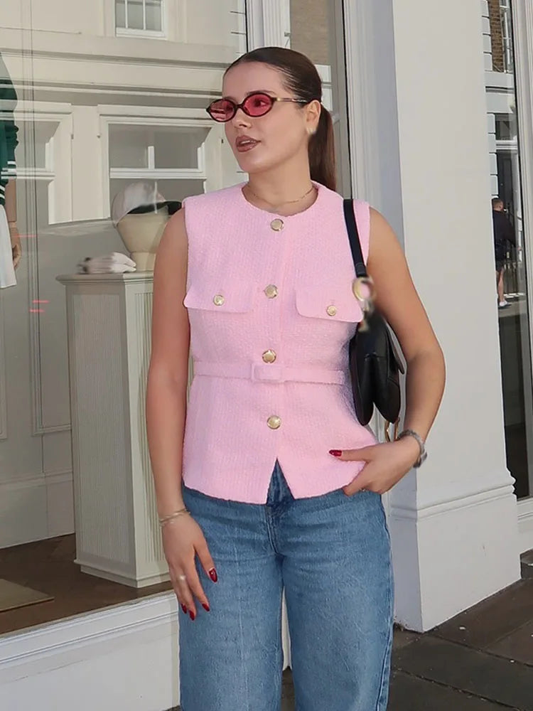 Woman wearing a pink sleeveless jacket with gold buttons, blue jeans, and sunglasses in front of a store.