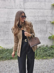 Woman in a beige coat holding a brown handbag against a concrete wall.