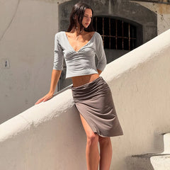 Woman in a light gray top and beige skirt standing against a textured wall.
