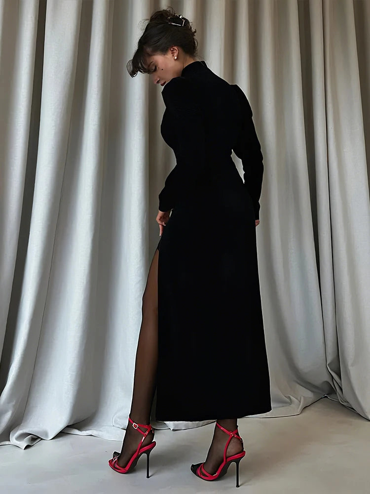 Woman in a black dress with a high slit standing against white curtains.