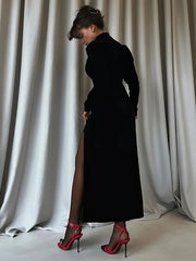 Woman in a black dress with a high slit standing against white curtains.