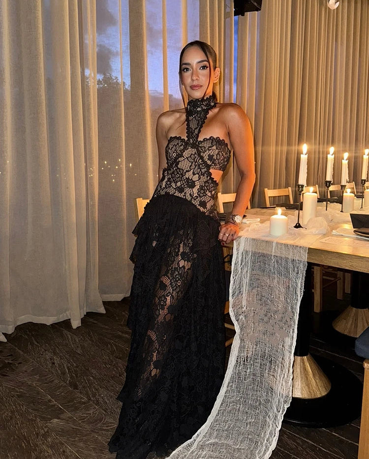 Woman in a black lace dress standing in a dining room with candles and curtains.
