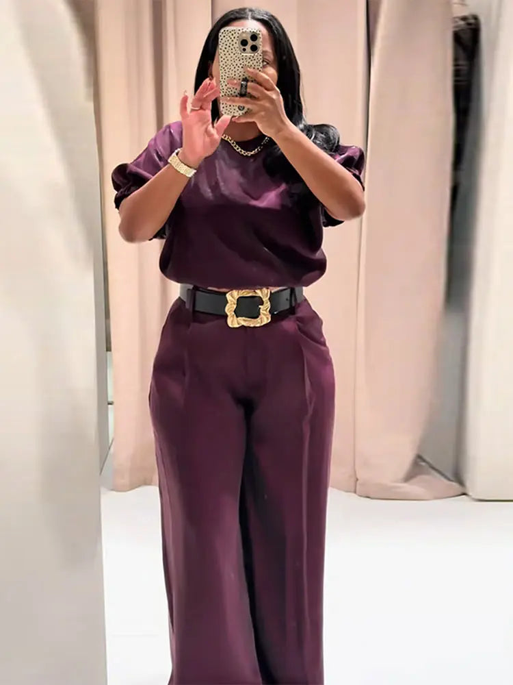Person wearing a purple outfit with a gold belt in front of beige curtains