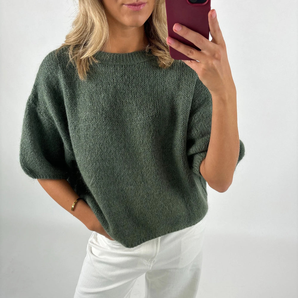 Person wearing a green sweater and white pants, taking a selfie.