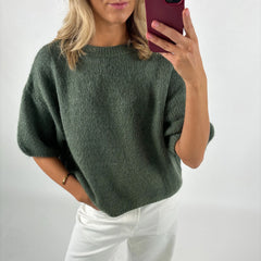 Person wearing a green sweater and white pants, taking a selfie.