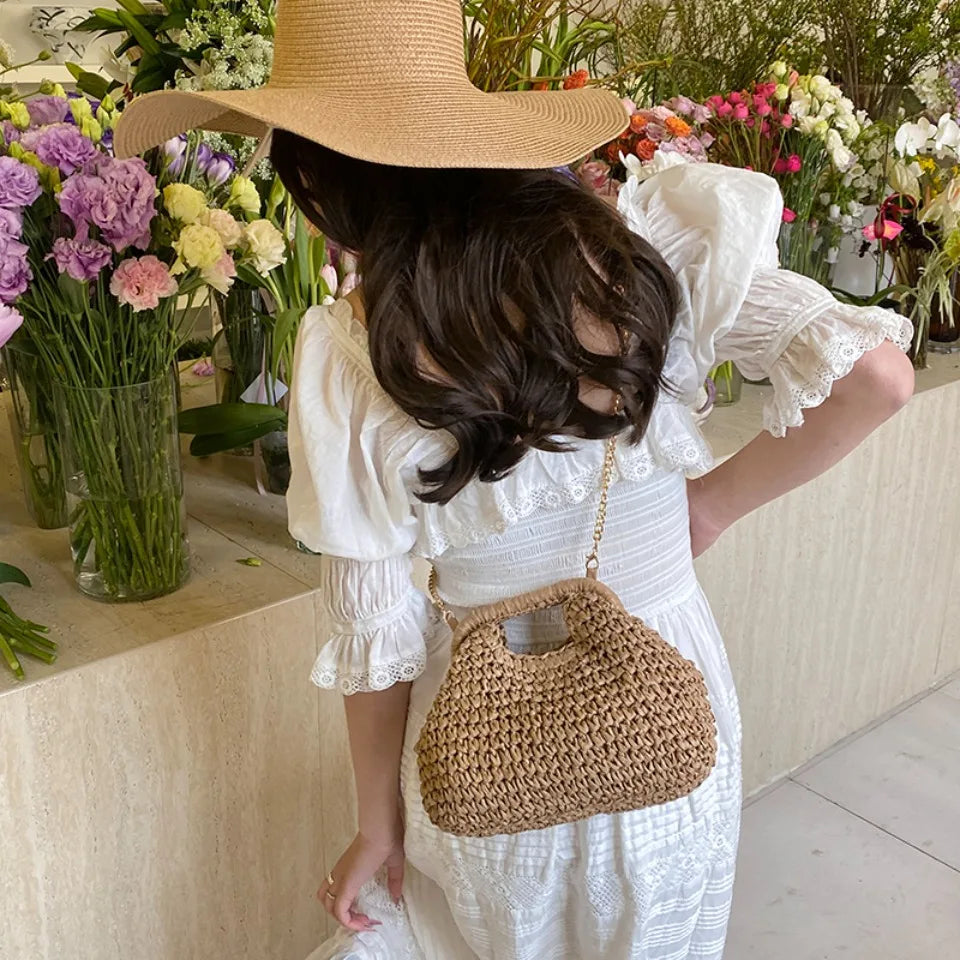 Woman in a white dress with a straw hat and bag standing among flowers.