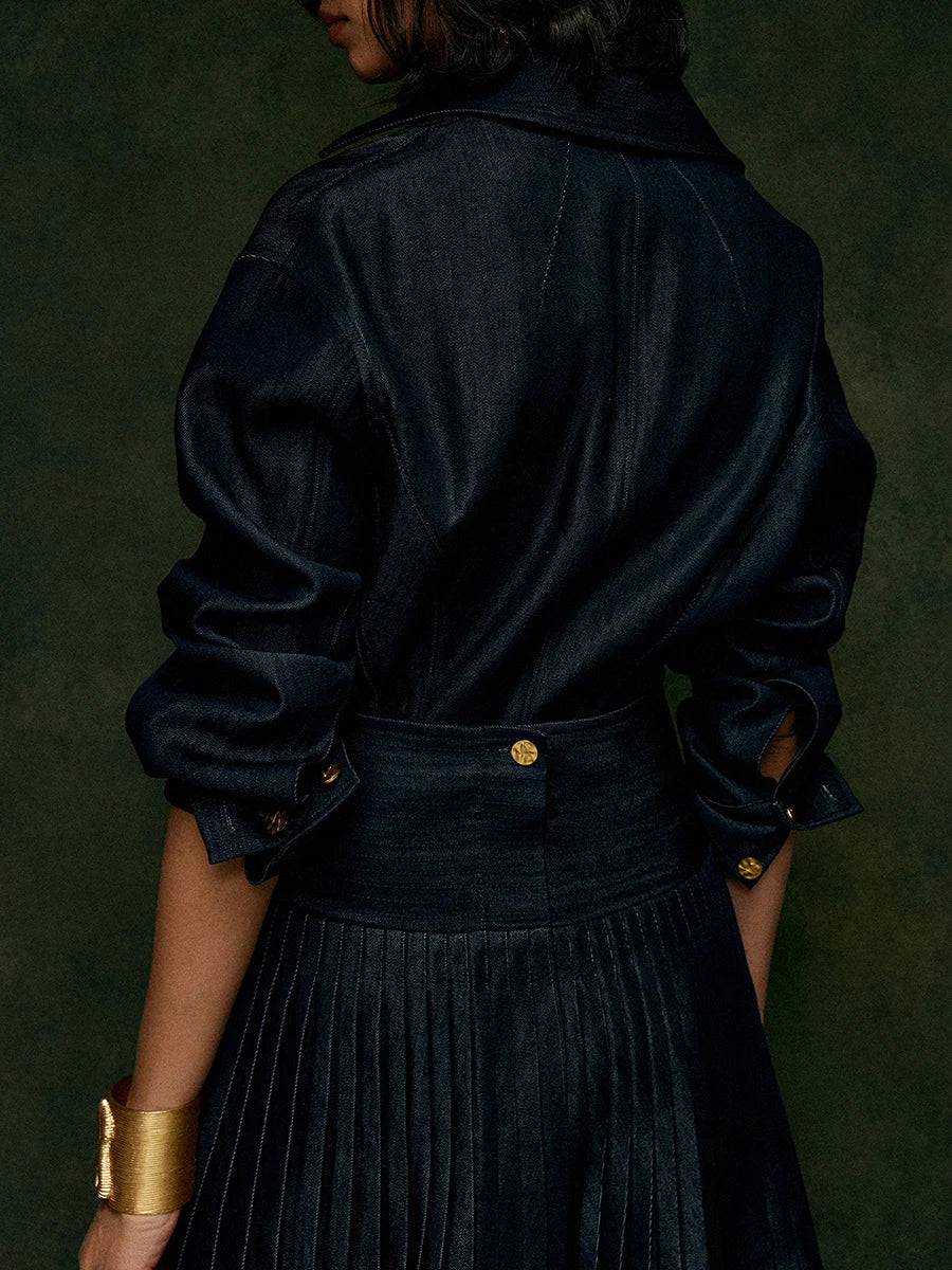 Person wearing a dark denim jacket and skirt with gold buttons against a green background