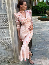 Woman in a light pink floral dress standing against a textured wall outdoors.