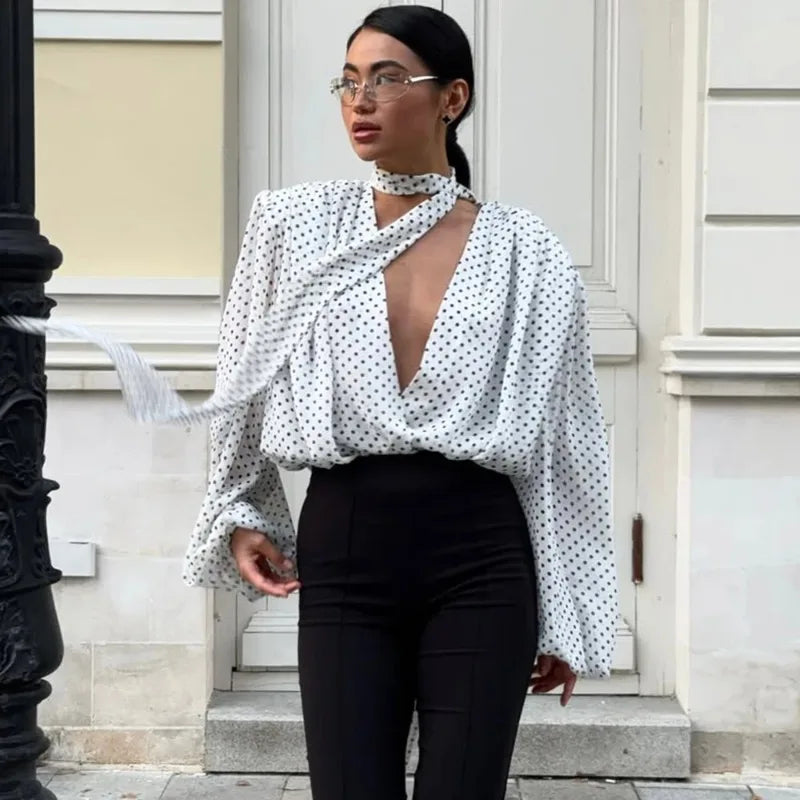 Woman wearing a white polka dot blouse with a deep neckline and black pants on a city street.