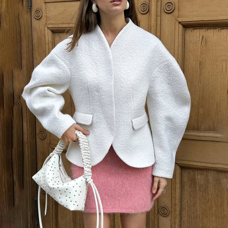 Woman wearing a white jacket and pink skirt holding a white handbag against a wooden door background