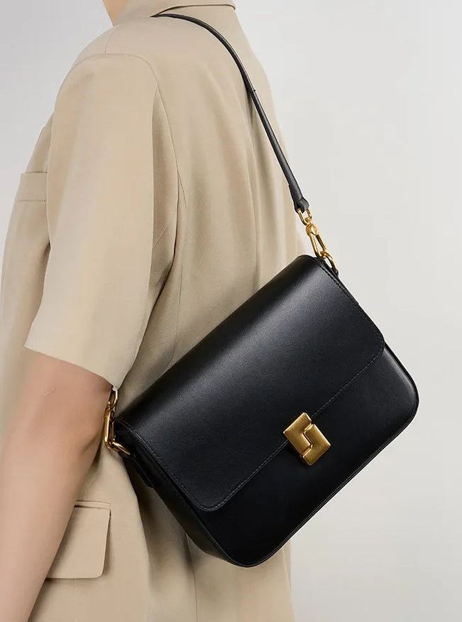 Black handbag with gold accents worn by a person in a beige outfit on a light gray background