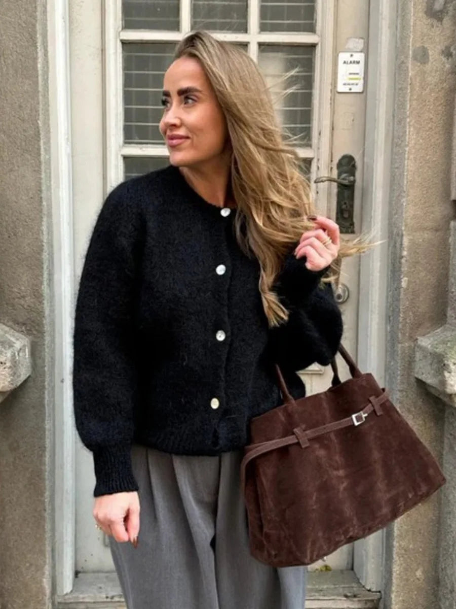 Woman wearing a black cardigan and holding a brown bag, standing outside a building.