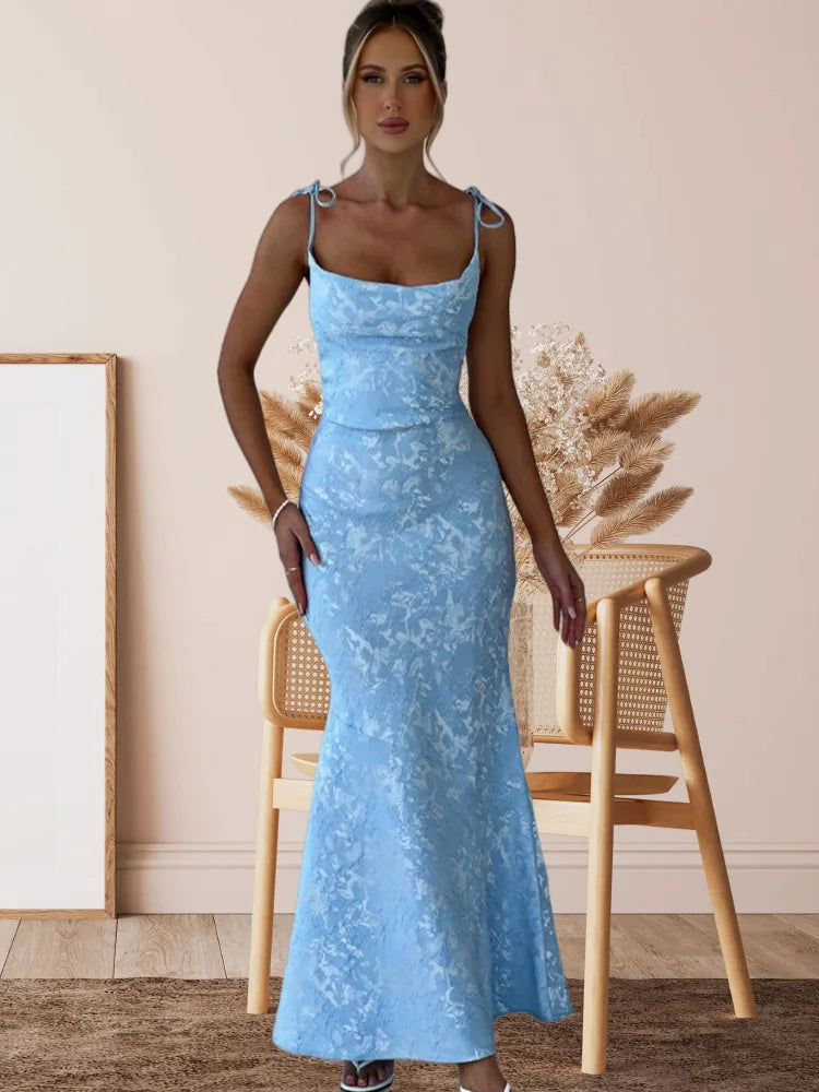 Woman wearing a blue lace dress standing in a room with a wooden chair and framed picture.